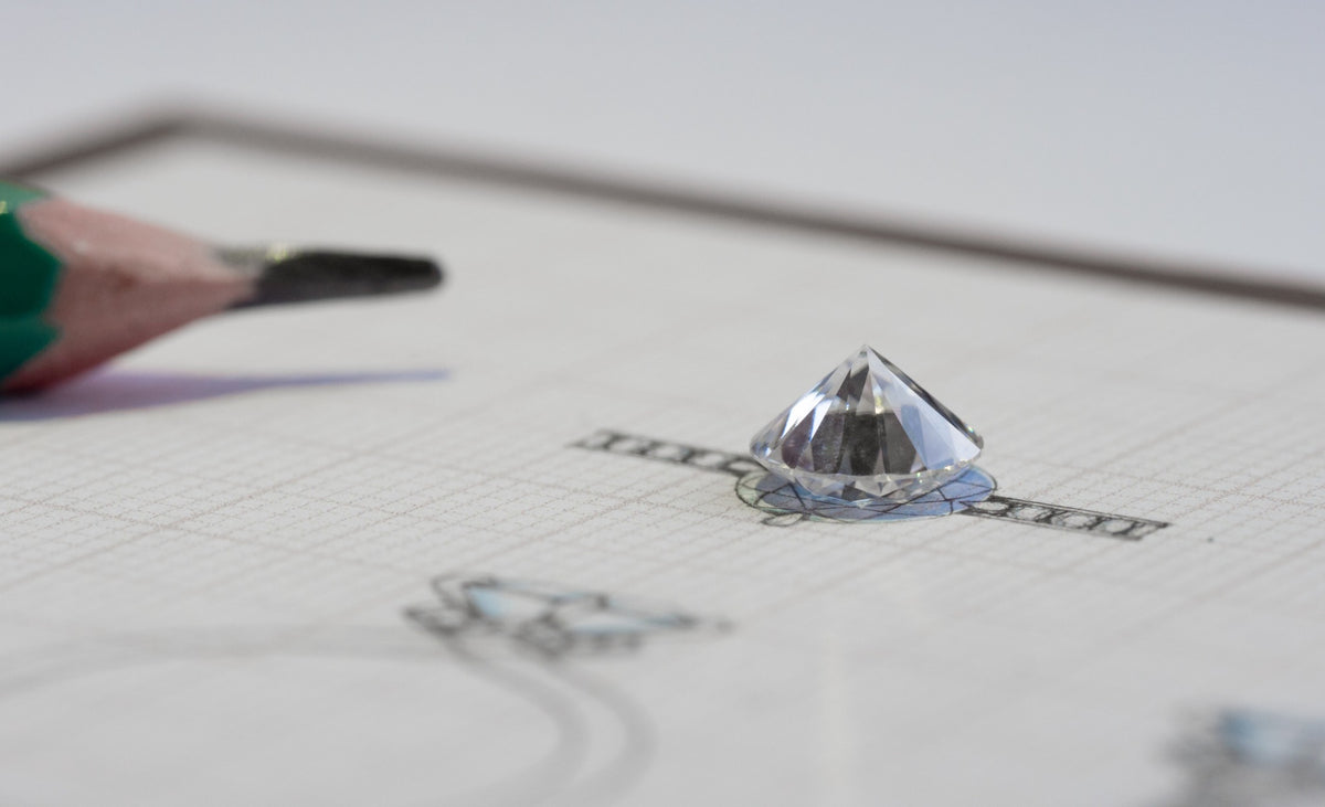 A loose diamond resting on a jewelry design sketch beside a pencil.